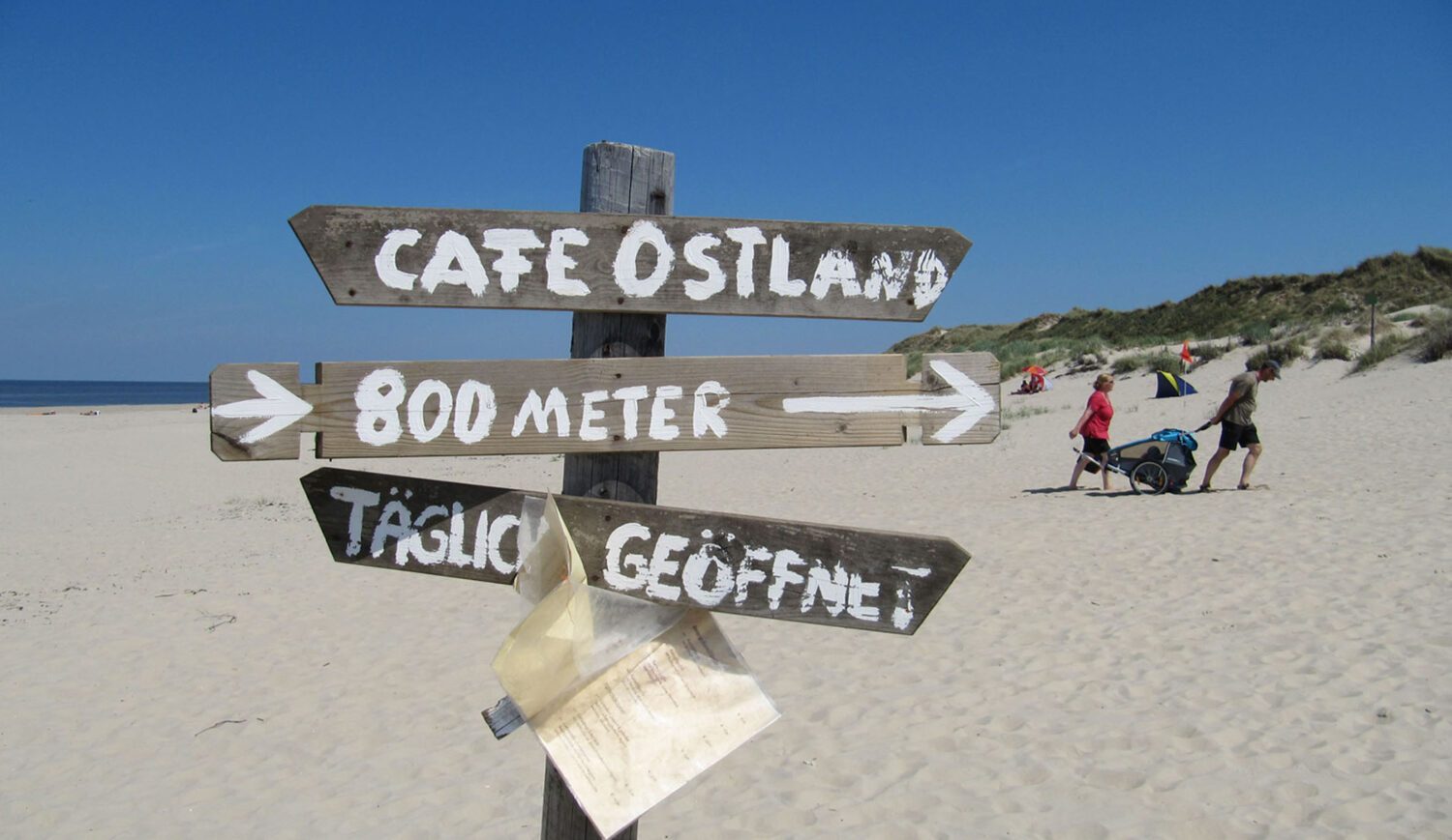 Dieses Schild am Strand von Borkum weist zu einem Café – damit gleich klar wird, ob der Weg sich lohnt, hängt die Speisekarte darunter